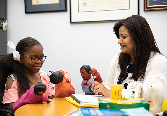 An elementary school student sitting at a table in an office looks down at two puppets held in her hands, and across from her is a smiling woman holding two other puppets. A notebook and folders are on the table.