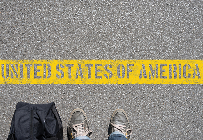 Two feet wearing shoes are pictured next to a black bag, at the bottom half of an image of a road with a thick yellow line on which “United States of America” is written.