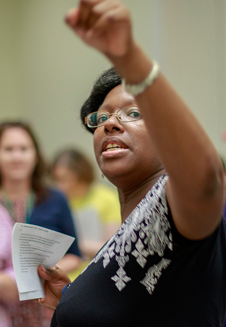 Adrienne Whaley holds notes in her one hand points towards something behind her as she teaches a room of individuals in the background.