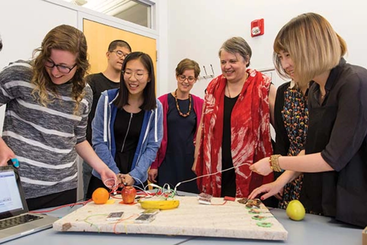 A smiling professor observing students working with a board which has articles including coins, fruit, and wires connecting to electronic devices.