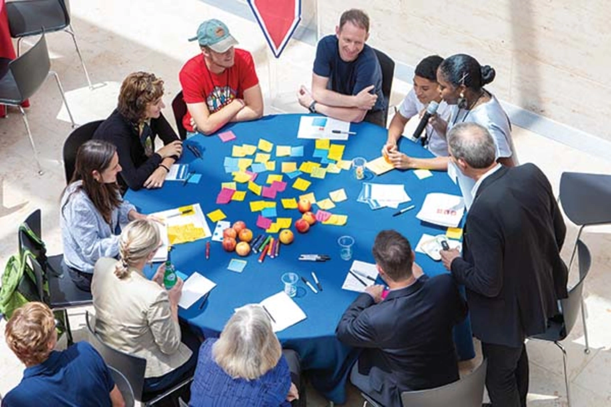 Several individuals at a round blue table. On the tabletop are colorful sticky notes, papers, pens, markers, and apples.