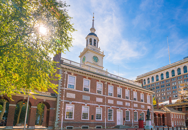 Independence Hall in Philadelphia appears against a blue sky, beside the sun shining behind a leafy tree