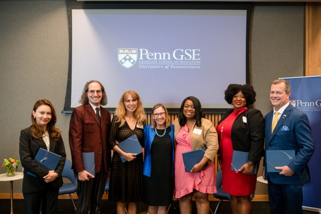 Dean Grossman stands in the middle of six smiling Education Alumni Award winners in front of a screen with the Penn GSE logo on it. 