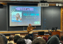 Felix Rosado, the program coordinator for Healing Futures with the Youth Art & Self-empowerment Project (YASP) and an adjunct professor at Chestnut Hill College, delivers a keynote to an audience of high school students in front of a projector screen with his headshot and name on it in a lecture hall.