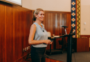 Penn GSE Senior Fellow Kandi Wiens stands by a podium with a microphone and laptop, holding a remote, smiling to an audience while giving a presentation on burnout.