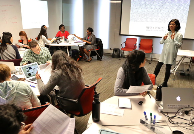 Students sit around four round tables as Ameena Ghaffar - Kucher leads the class with a screen behind her with a projection on it that reads “Pause and Reflect.”