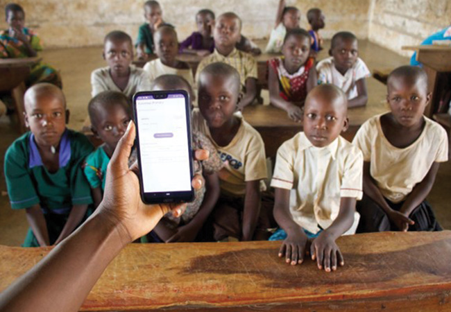 A hand extends forward holding a smartphone with the Skizaa app on the screen in front of a classroom full of elementary school children in Uganda's Mayuge district.