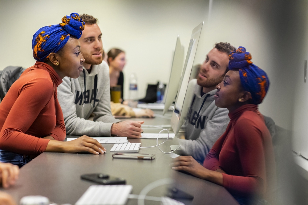 Female and male students look at a computer together. You see their reflections on the right half of the image.