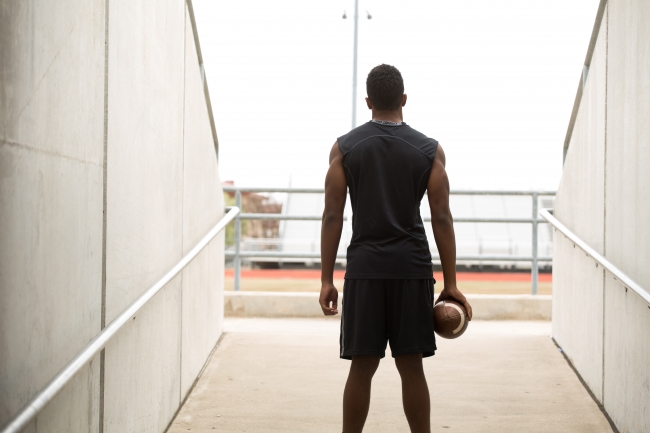 A teen in gym clothes holding a football stands in the entrance to a stadium looking out onto the field.