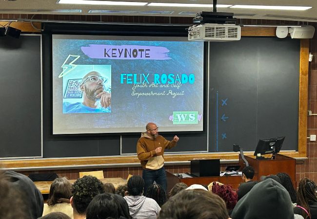 Felix Rosado, the program coordinator for Healing Futures with the Youth Art & Self-empowerment Project (YASP) and an adjunct professor at Chestnut Hill College, delivers a keynote to an audience of high school students in front of a projector screen with his headshot and name on it in a lecture hall.