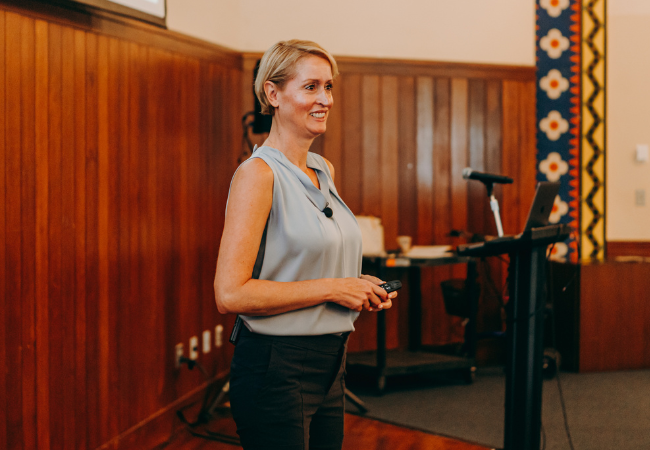 Penn GSE Senior Fellow Kandi Wiens stands by a podium with a microphone and laptop, holding a remote, smiling to an audience while giving a presentation on burnout.