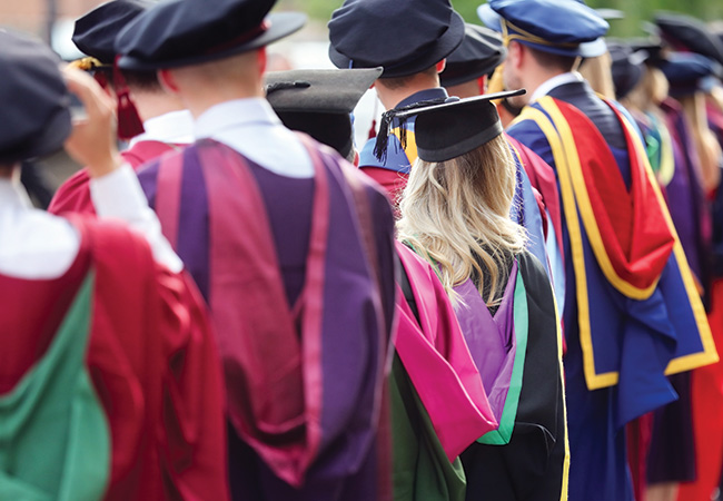 University faculty and leadership wearing commencement regalia including hoods in many colors stand in a row with their backs to the camera