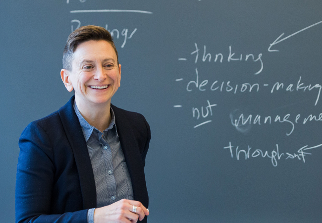 Abby Reisman smiles during class, standing in front of a chalkboard and holding apiece of chalk.