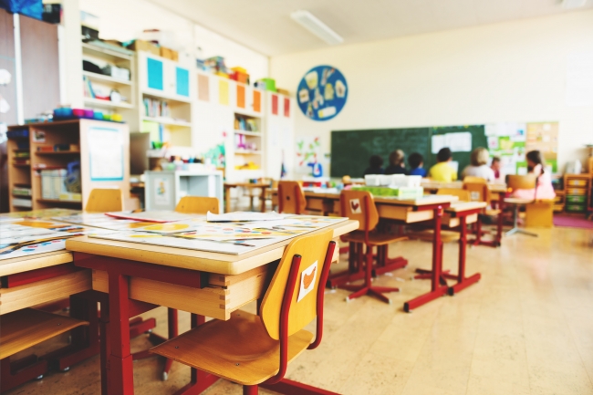 A cluster of wooden student desks in a bright, sun-filled elementary school classroom. In the background, out of focus, appears to be a reading circle. Credit: Adobe Stock.