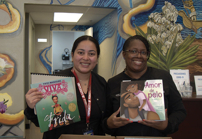 Two volunteers stand in the Puentes de Salud medical clinic with a mural painted on the wall behind them, holding children’s books in Spanish donated by A Book a Day, a non-profit run by Penn GSE student Sibylla Shekerdjiska-Benatova