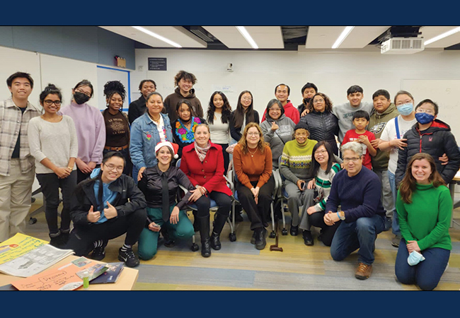 A large multicultural group of 27 youth, parents, and other adults pose together in a semicircle, smiling at the camera in a classroom.