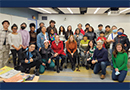 A large multicultural group of 27 youth, parents, and other adults pose together in a semicircle, smiling at the camera in a classroom.