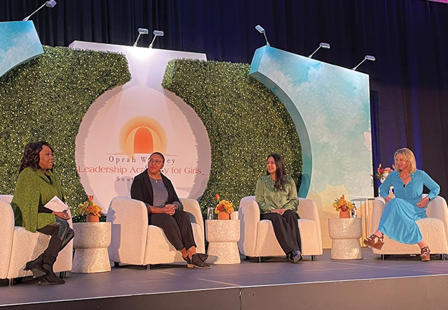 Four women sit in upholstered chairs on a black stage, a backdrop set piece behind them of walls painted blue with white clouds and greenery in the shape of an “O” around the Oprah Winfrey Leadership Academy for Girls logo. From left to right are the panel moderator in a green shawl, Charlotte Jacobs in a gray shirt with a black shawl, a participant in a green satin button-down shirt, and a participant in a light blue dress, discussing trauma-informed practice in education.