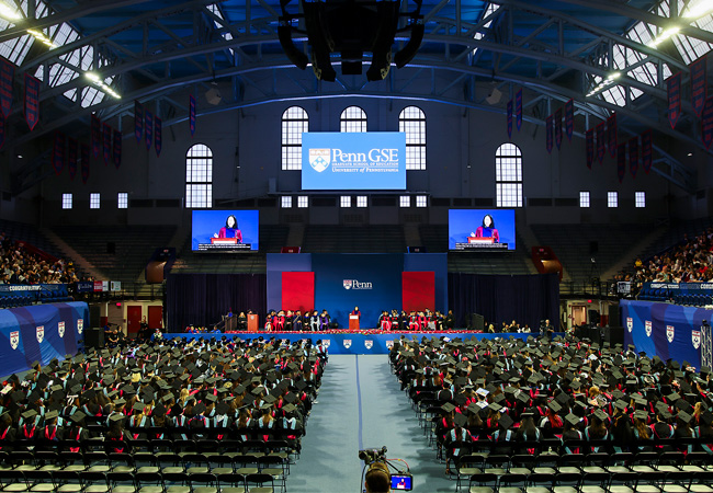Penn GSE University of Pennsylvania, with graduates seated in the Palestra under banners, facing a stage where a Angela Duckworth is visible on two large screens.