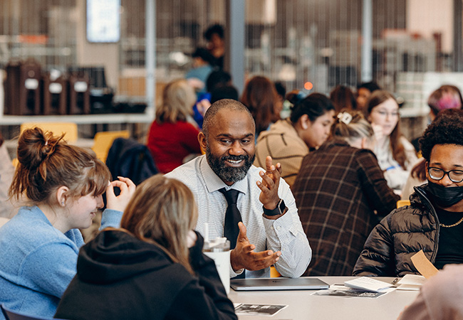 A teacher – a Black man with a trimmed beard in a white shirt and tie – sits facing the camera. He is at a table during a breakout session. He is smiling animatedly and speaking with a small, diverse group of students. Similar groups working at other tables are slightly out of focus in the background.