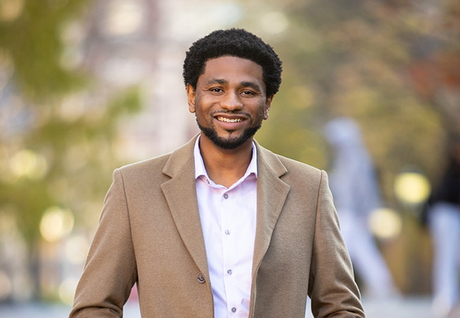 Portrait of Damani White-Lewis outdoors, smiling, with a green leafy background. He is wearing a tan sports jacket.