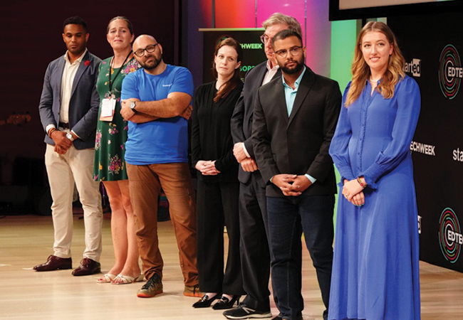 A diverse group of 7 educational entrepreneurs dressed in business casual clothes stand lined up on stage awaiting the results of a competition.