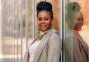 Ericka S. Weathers, with a warm smile, stands confidently, her braided hair elegantly styled into an updo, complemented by large hoop earrings, a vibrant beaded necklace, and a chic beige blazer, reflected against a glass surface.