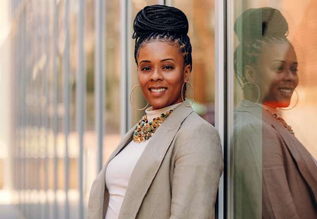Ericka S. Weathers, with a warm smile, stands confidently, her braided hair elegantly styled into an updo, complemented by large hoop earrings, a vibrant beaded necklace, and a chic beige blazer, reflected against a glass surface.
