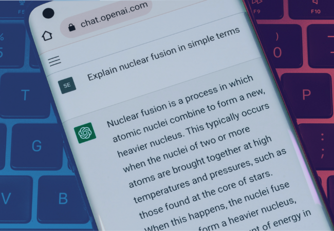A smartphone is held in the center of the image with the ChatGPT website open on its browser. The phone is above a computer keyboard. You can see that the person has input “explain nuclear fusion in simple terms” as their prompt and the AI is generating an answer in return, illustrating how the program works for end-users.