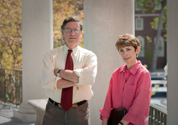 Man with glasses standing and woman with short hair sitting outside.