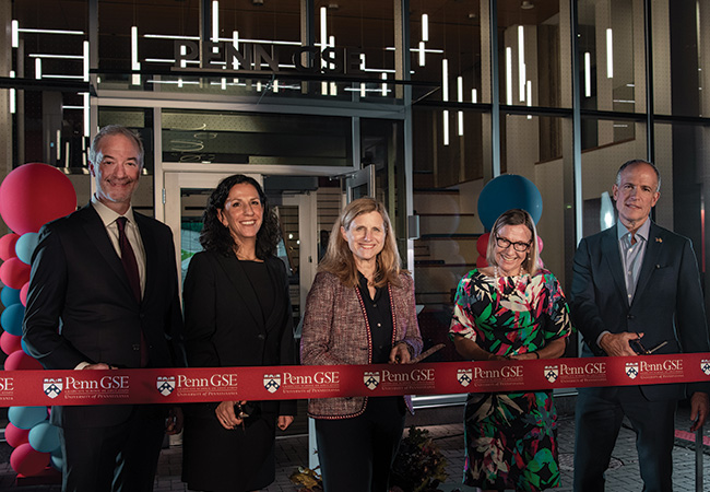 From left, GSE Board of Advisors chair Jeff McKibben, GSE Dean Katharine Strunk, Penn President Liz Magill, former GSE Dean Pam Grossman, and GSE board member and former chair Doug Korn cut a red “Penn GSE”-branded ribbon with scissors in front of the glass façade of the new building entrance at night.
