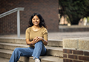 Penn GSE student Meresa García sits outside on cement steps in front of a brick wall on the University of Pennsylvania campus wearing a tan t-shirt and blue jeans, smiling at the camera