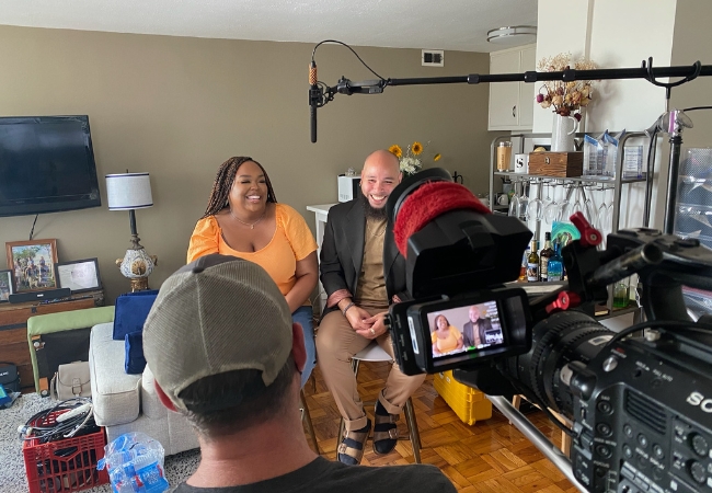 Penn GSE alums Brittany Robertson and Serrano Legrand sit in a living room in front of a camera and boom mic while a House Hunters crew member interviews them for an episode of the show
