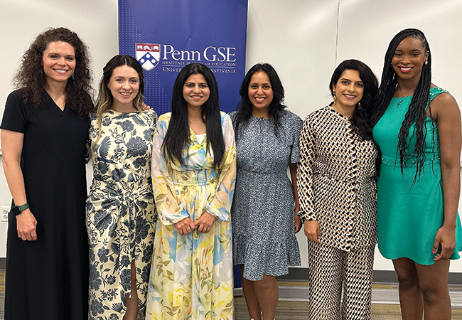 The six women in the inaugural cohort of Jacobs Fellows stand, smiling, in a classroom in front of a Penn GSE banner.