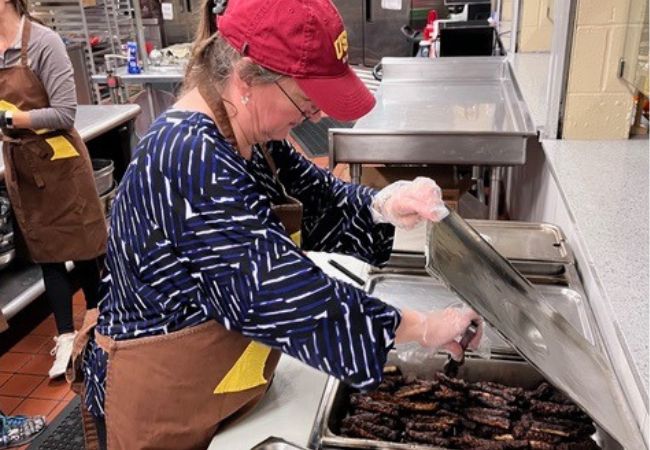Julee Gard, wearing a red cap, a navy blue and white striped shirt, and a brown apron, is focused on serving sausages at a soup kitchen in Joliet, Illinois, alongside her University of St. Francis colleagues.