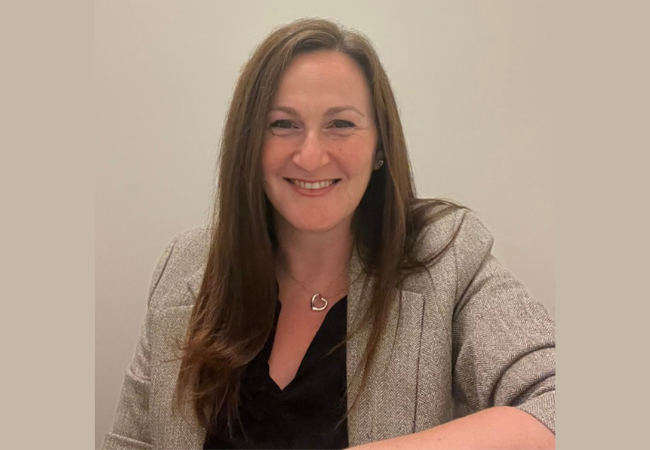 A headshot portrait of Lara wearing a herringbone blazer and smiling against a neutral background. 