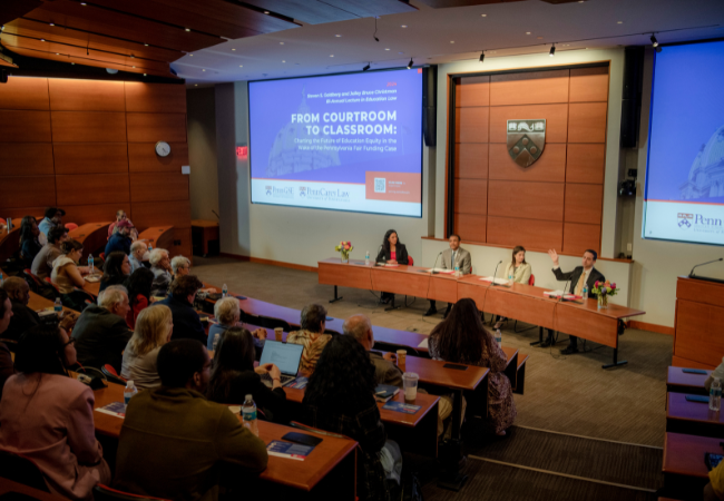 A view of the three panelists and Dean Katharine Strunk sitting at a table in front of the packed lecture hall under a screen that reads, “From Courtroom to Classroom.”