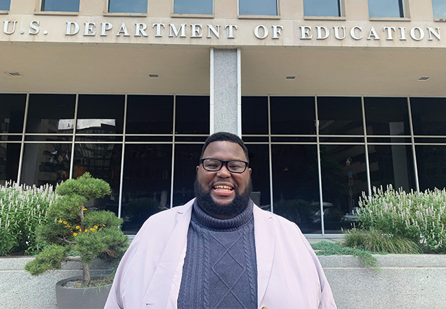 Mark Ziegler-Thayer smiles at the camera wearing a white blazer over a gray sweater, standing in front of the Lyndon Baines Johnson Department of Education Building in Washington, D.C. The words “U.S. Department of Education” are mounted on the face of the building in large metal letters above Mark’s head.