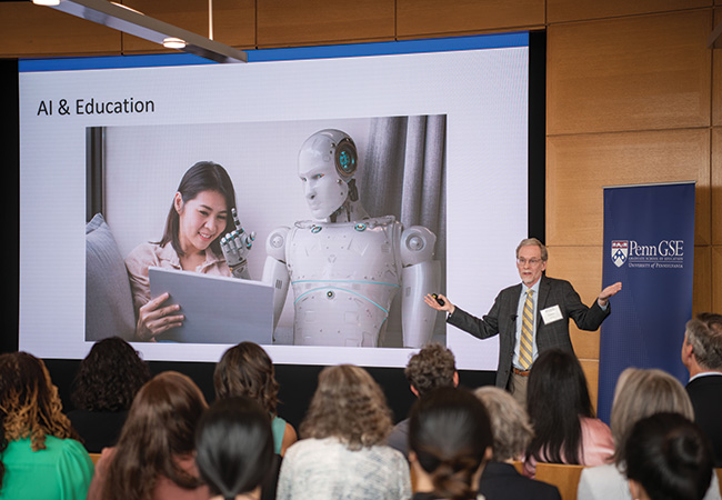 Dr. James Lester, Distinguished University Professor of Computer Science and Director of the Center for Educational Informatics at North Carolina State University, stands with his arms spread, delivering the keynote address to a seated crowd at the launch of Penn GSE's McGraw Center for Educational Leadership. On the screen behind him is an image of a robot providing instruction to a woman as they view a tablet together under the label "AI & Education," the topic of Dr. Lester's talk.