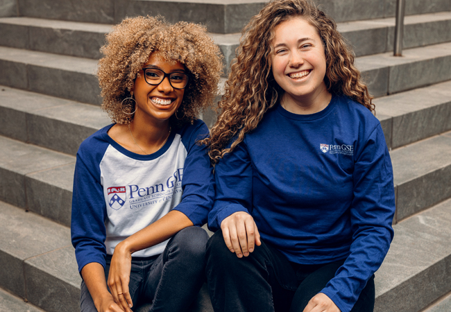 Two graduate students sit on the stairs outside Penn GSE in blue GSE long-sleeved t-shirts.