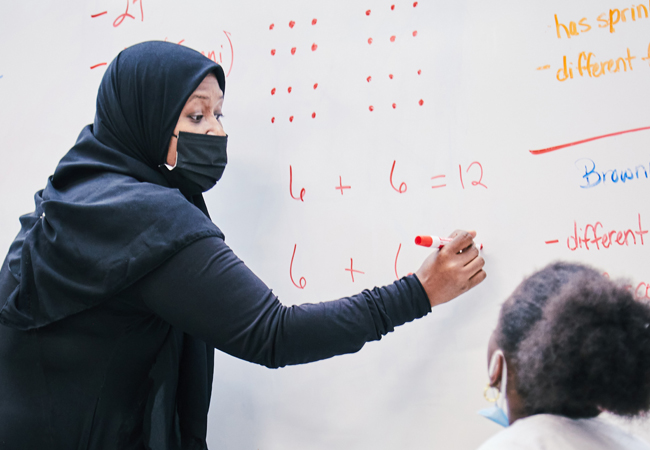 A teacher wearing a mask does arithmetic on a white board with a student.