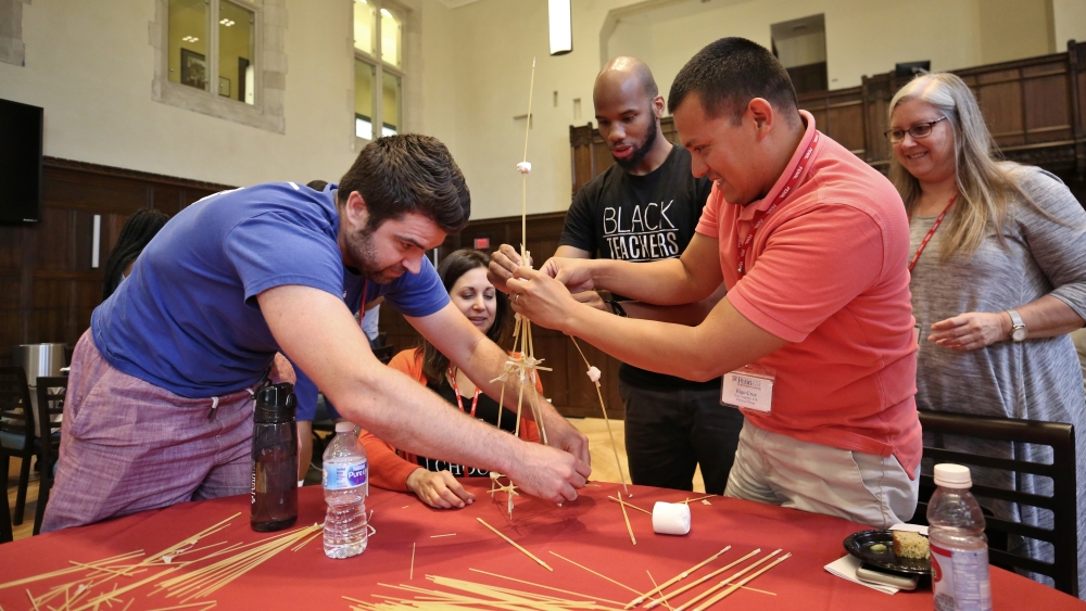 Several people build a structure out of sticks and marshmallows top of a table