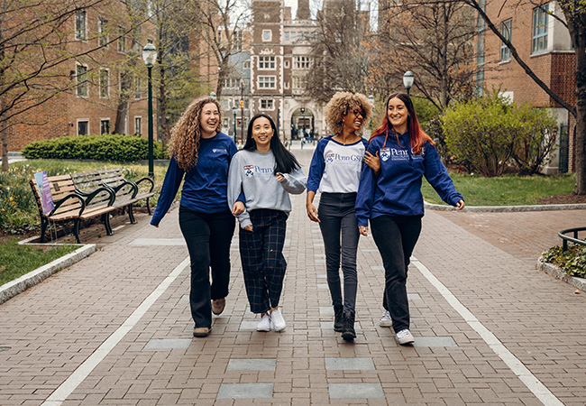 Four Penn GSE students wearing Penn GSE sweatshirts in blue, gray, or white walk down a path on the Penn campus. They are smiling and laughing.