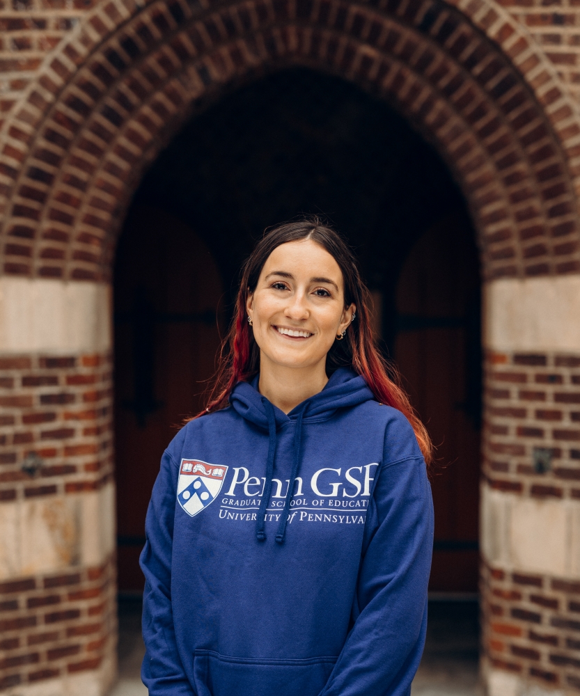 Female graduate student wearing a dark blue Penn GSE hooded sweatshirt with a brick arch on the Penn campus in the background.