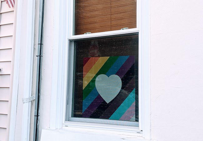 A sign with a heart over a pride flag hangs in the window of a house. 