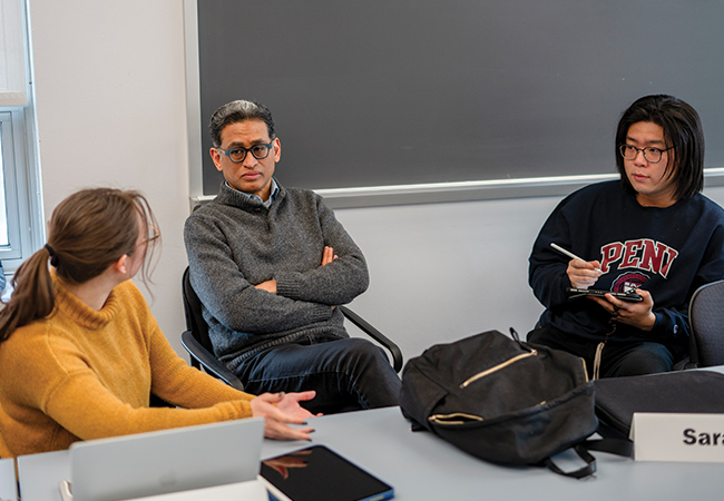 Rand Quinn sits at a table with his arms crossed engaged in discussion with two students