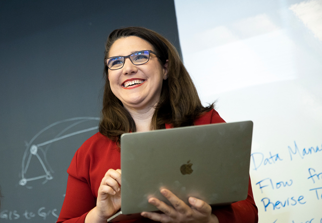 Sarah Schneider Kavanagh smiles and holds her laptop while standing in front of achalkboard with a diagram drawn on it.