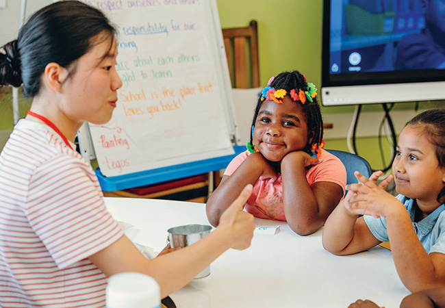 A young woman, a member of a classroom instruction team wearing a red-and-white striped shirt and holding a tin cup in her left hand with her right thumb up, sits on the left across a table from three students paying close attention to her instruction. The leftmost student sits in the center of the frame resting her elbows on the table, hands propping up her head, smiling at the camera with bright multicolored flower-shaped beads in her hair.