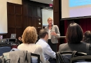 Penn GSE Senior Fellow Kandi Wiens stands wearing a gray blazer in front of a group of seated school district leaders from around the Philadelphia region delivering a presentation on burnout in a wood-paneled room with a screen showing a slide behind her at a Center for School Study Councils meeting