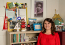 Nimet Eren, dressed in red attire, stands in her office surrounded by a collection of artifacts that reflect her journey as an educator.